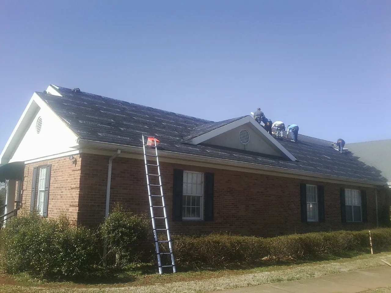 Crew tearing shingles off a brick ranch roof with a ladder set — Metal Roof Repair work by our crew in Fertile