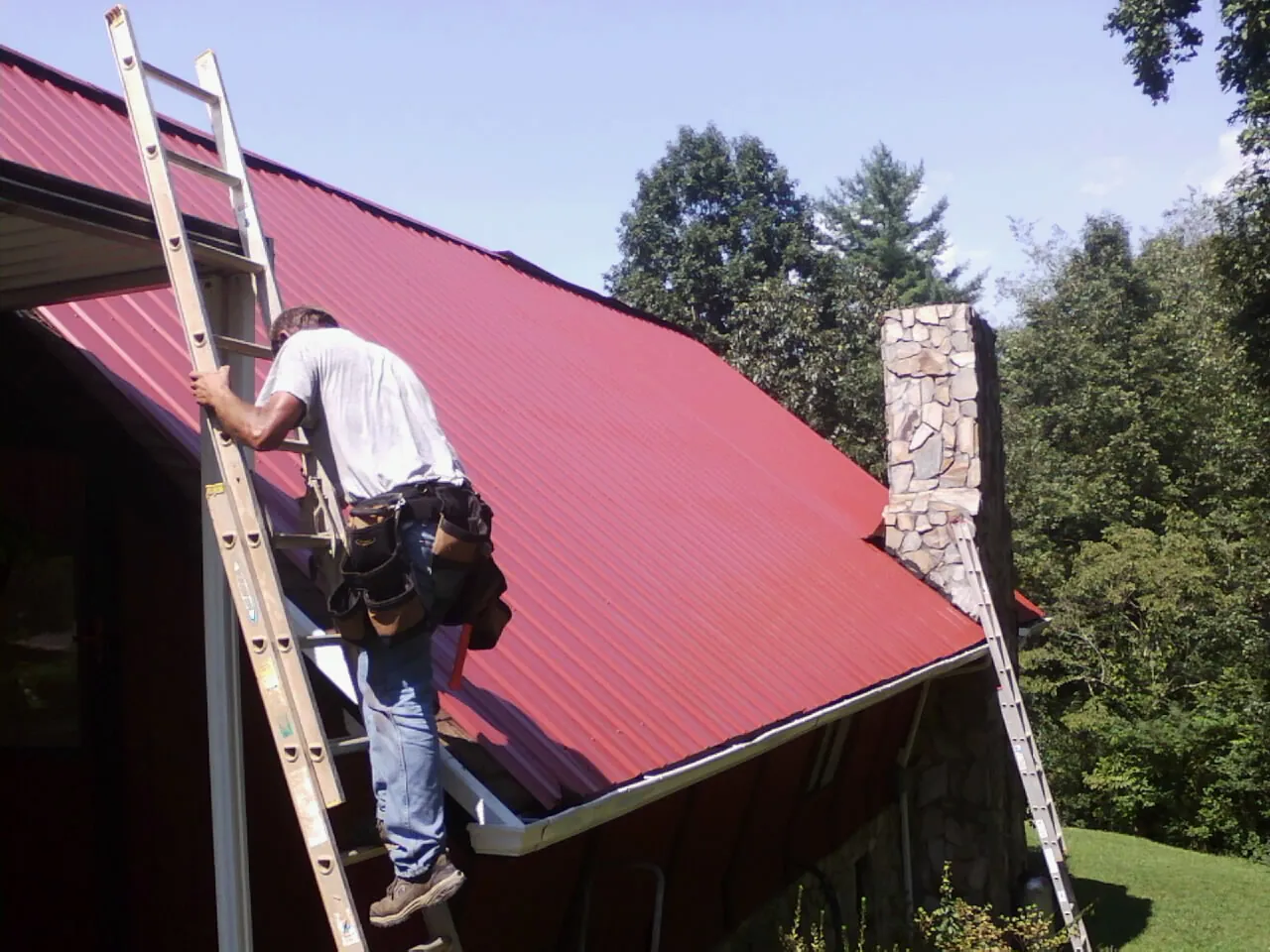 Roofer on a ladder beside a red metal roof and stone chimney — Soffit & Fascia Repair by Roofing Brothers in Fertile